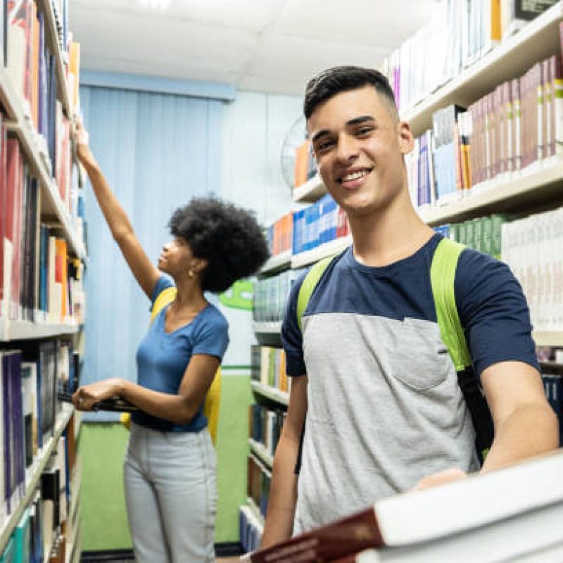 stu img Portrait of a teenager boy carrying some books in a library with colleage in the background