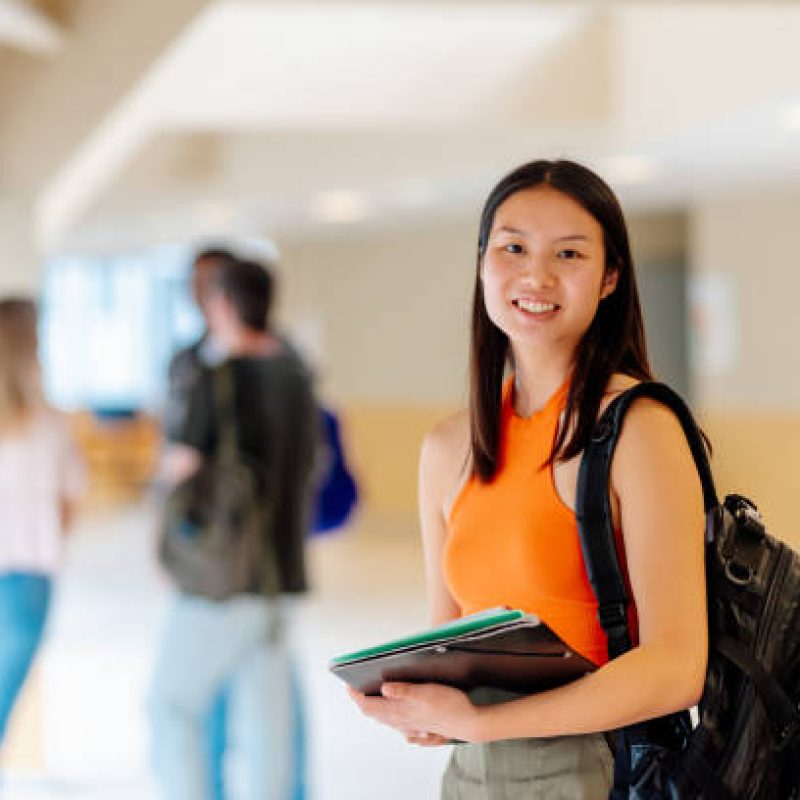 istockphoto-1759509366-612×612 Portrait of an Asian girl with a backpack and school supplies in the hallways of the university campus with her classmates in the background.
